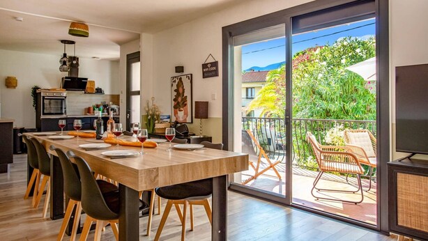 A modern kitchen with a dining area, large sliding glass doors to a balcony, and a view of a lush garden. - Villa Du Soleil by Villa Plus (Laroque-des-Albères)