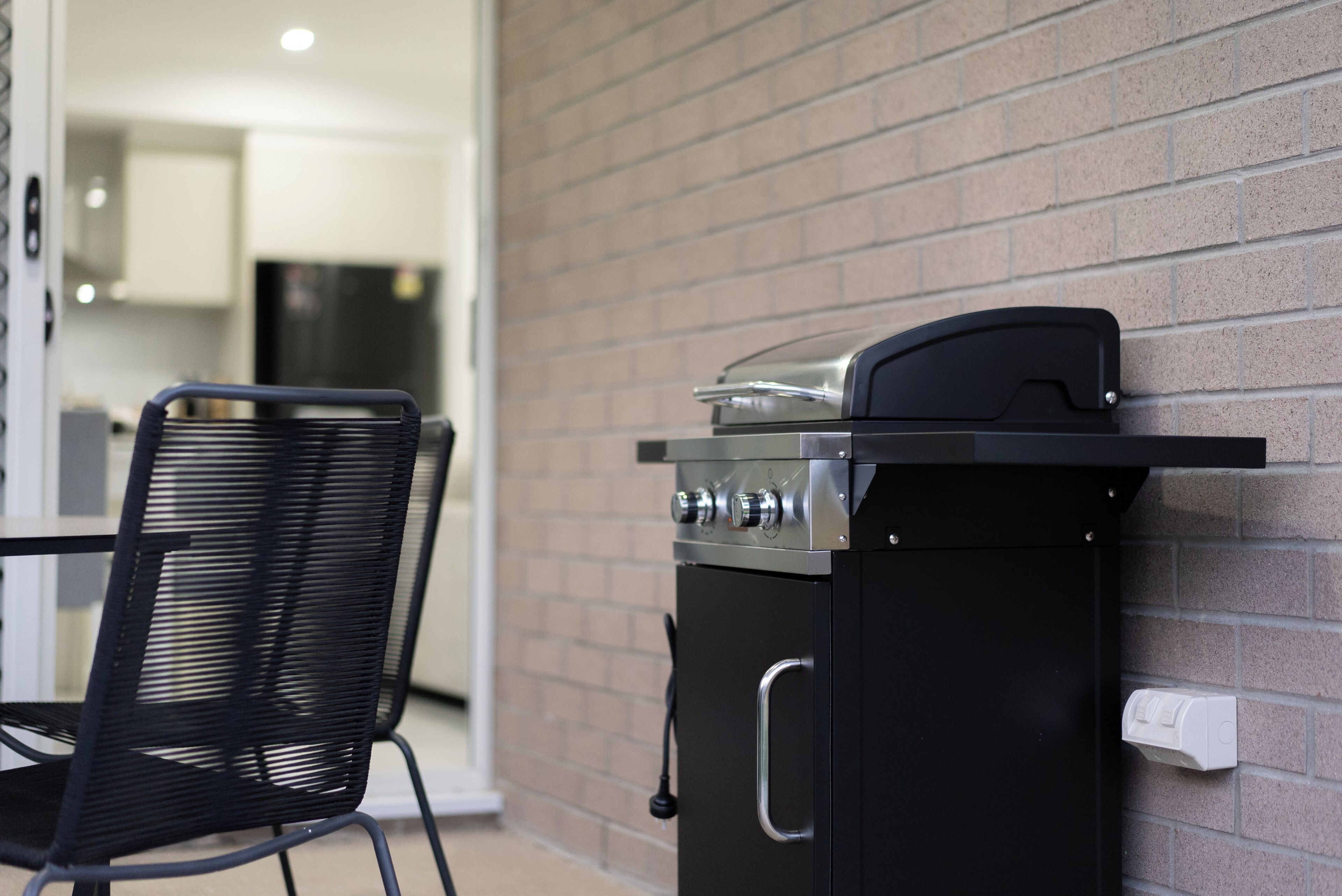 A black barbecue grill attached to a brick wall, a black metal chair, and a glimpse of a kitchen area in the background.