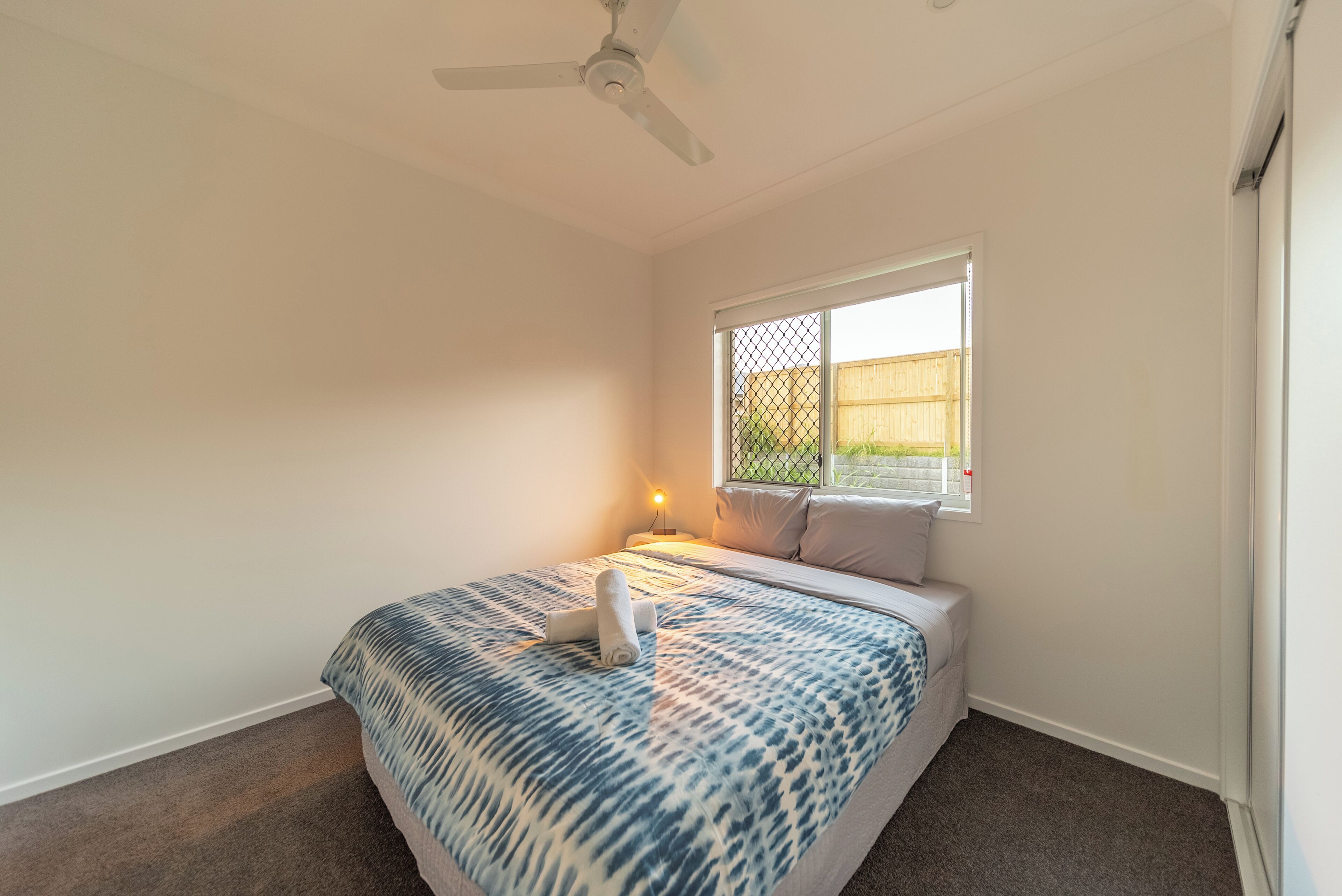 Ein Schlafzimmer mit einem Bett, einem Deckenventilator, einem Fenster mit Blick auf einen Zaun und einem Nachttisch mit Lampe.
