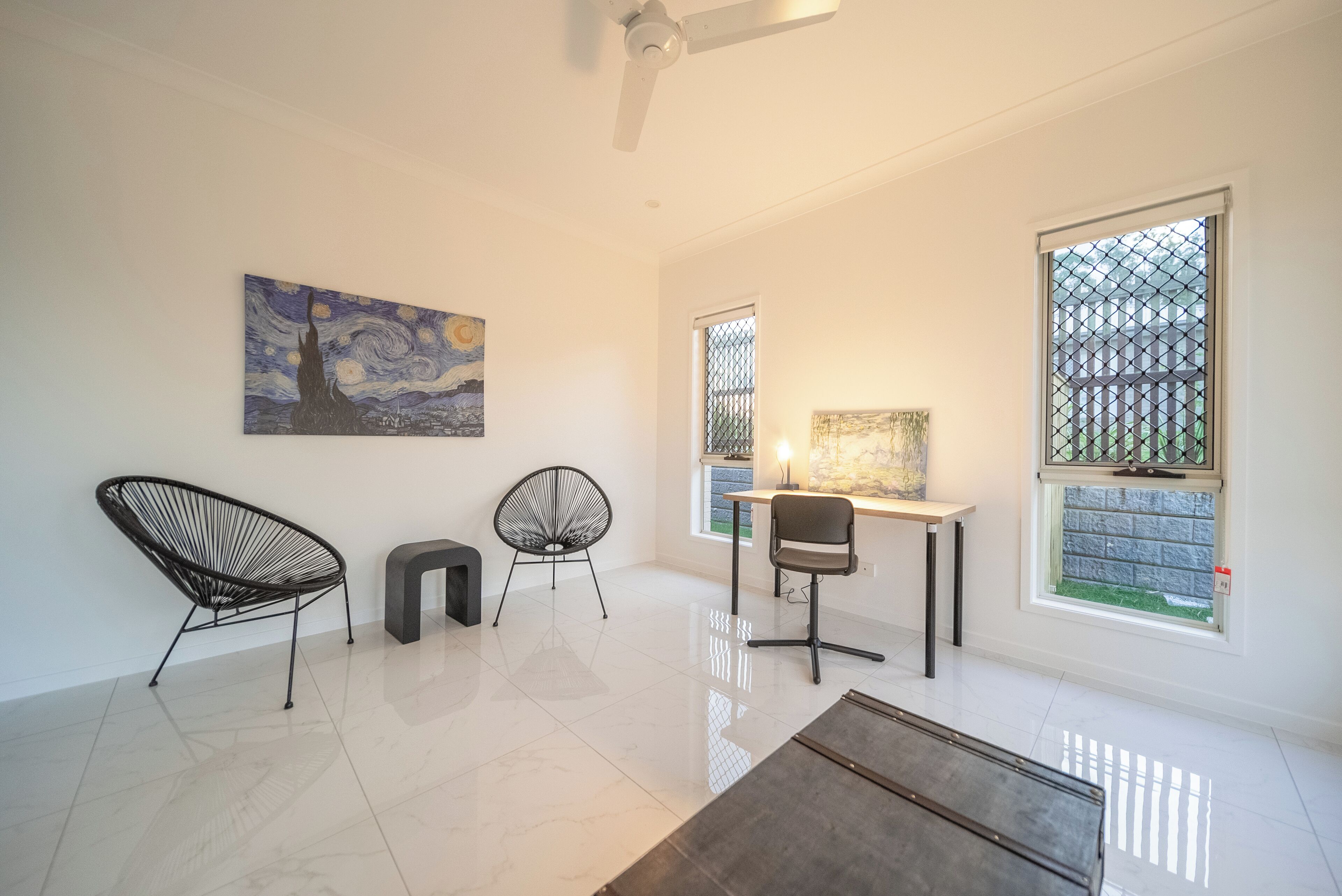 Un salon moderne avec un ventilateur de plafond, un tableau, deux fauteuils, un bureau et de grandes fenêtres.