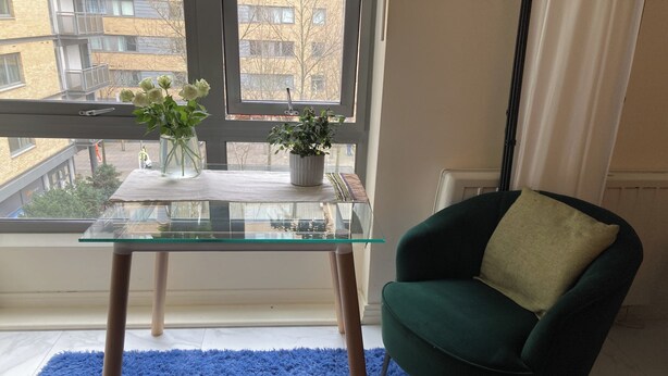 A room with a glass-top table, a green armchair, a blue rug, and a window with a view of buildings.