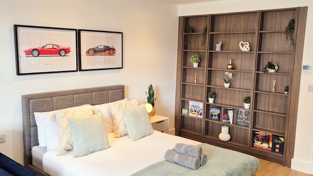 Een moderne slaapkamer met een groot bed, een houten hoofdbord, een nachtkastje en een ingebouwde boekenkast met diverse spullen.