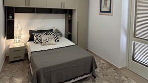 Een slaapkamer met een bed, nachtkastje, lamp, kledingkast en een raam met jaloezieën.