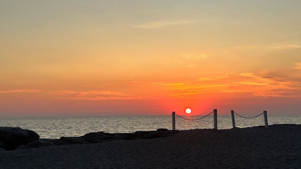 Een zonsondergang boven een strand, met een hek van gaas in de voorgrond.