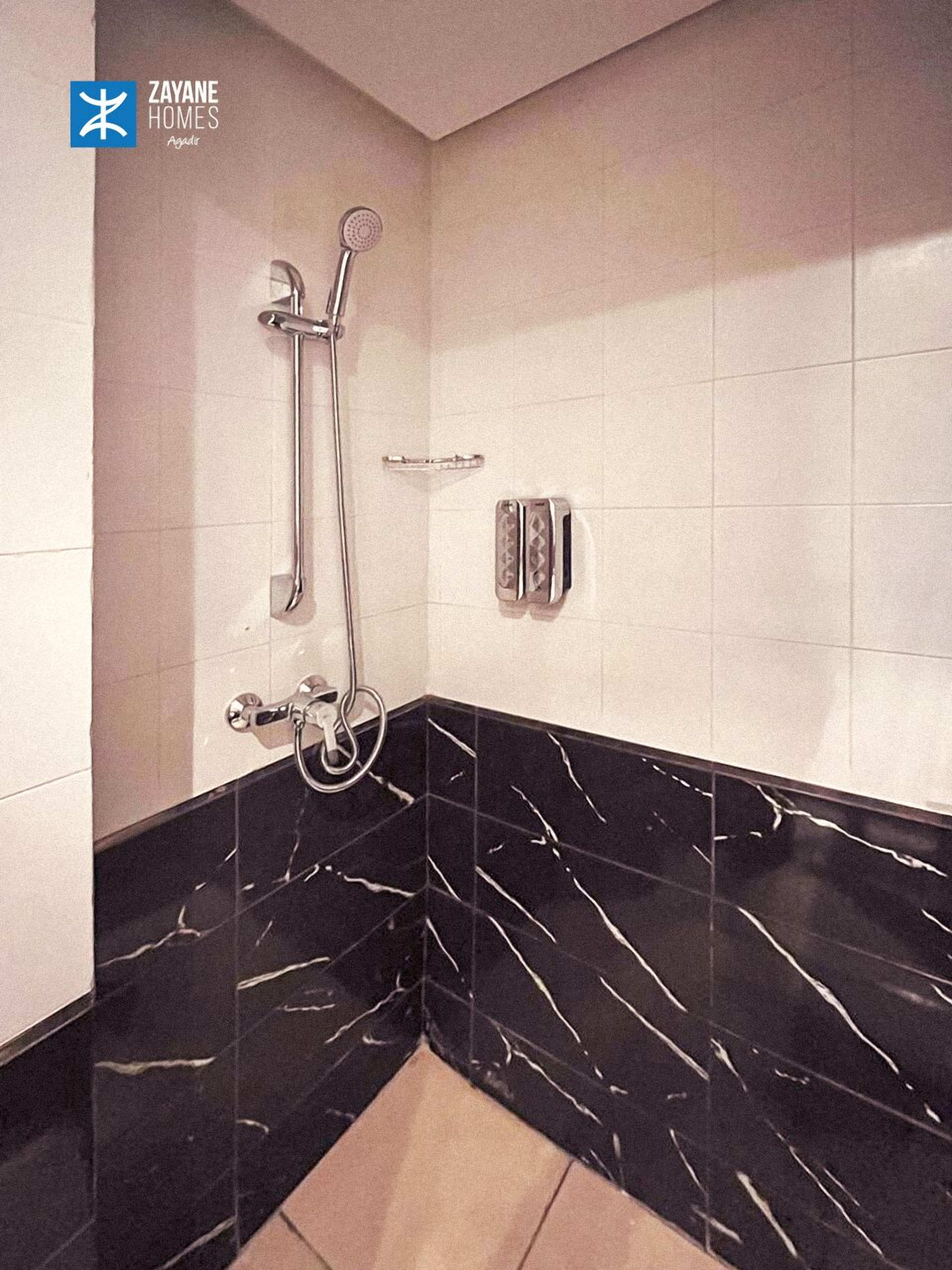 Ein modernes Badezimmer mit Waschtisch aus schwarzem Marmor, einer Dusche mit Handbrause und einer wandmontierten Bedienkonsole.