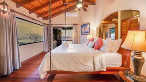 A spacious bedroom with a large bed, wooden ceiling beams, a ceiling fan, and a view through a large window. - Enchanting 6 Bedroom Villa with Incredible Views- Fully Staffed -Walk to Beach (Tamarindo)