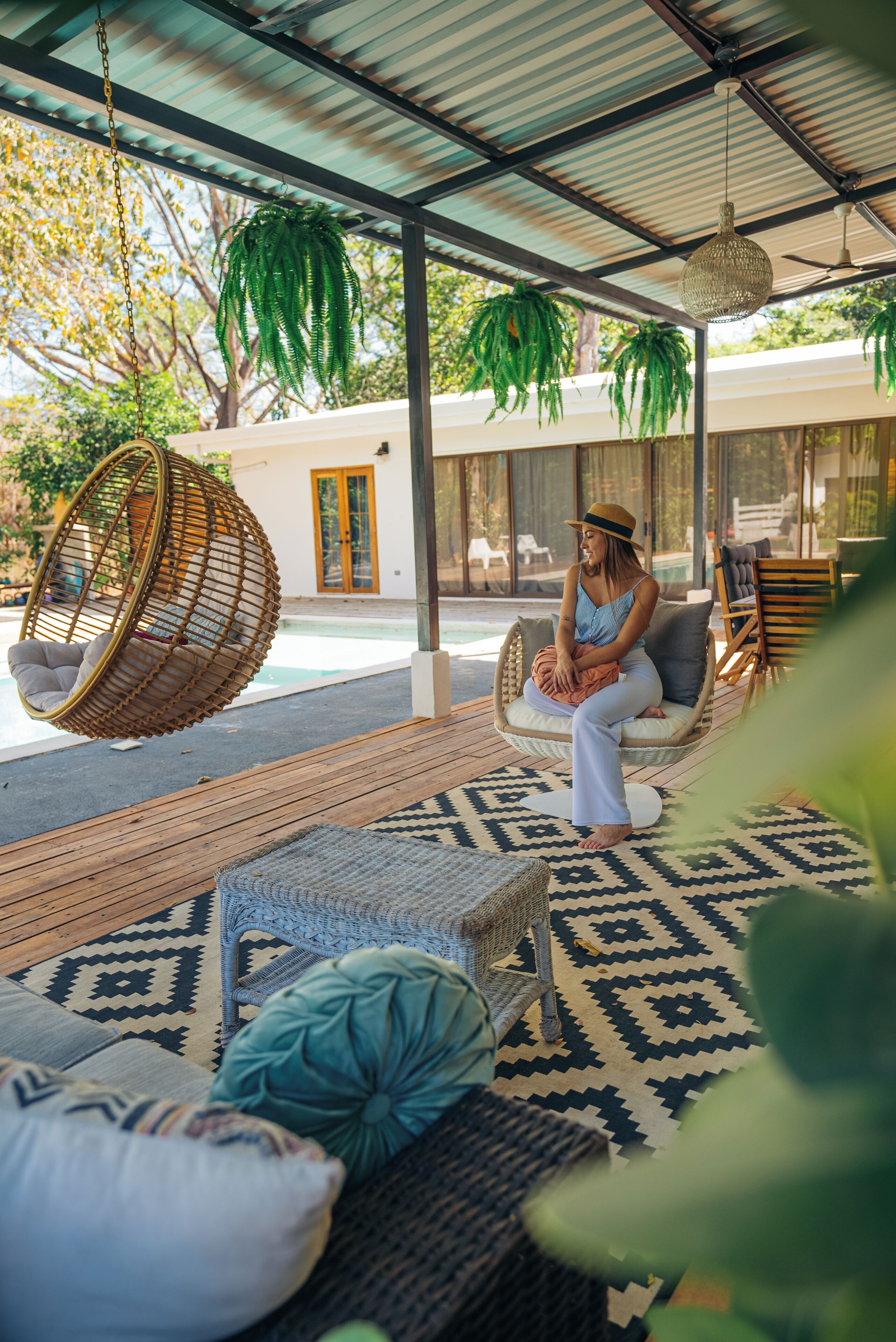 Un espacio de estar al aire libre techado con sillas de mimbre y una silla colgante, una alfombra estampada y una vista a una piscina y una casa al fondo.