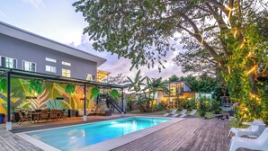 Ein moderner Poolbereich im Freien mit einer Holzterrasse, Liegestühlen und einer überdachten Sitzecke mit farbenfrohem Wanddesign.