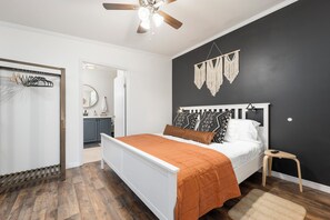 Een slaapkamer met een bed, een klein krukje, een plafondventilator, een macramé wanddecoratie en een houten vloer.