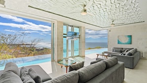 A modern living room with a pool view, grey sofas, and a ceiling with a wavy pattern. - Ultimate Luxury Villa: Hot Tub, Gourmet Kitchen & Stunning Ocean Views - Fully Staffed (Tamarindo)