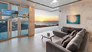 A modern living room with a large sofa, a coffee table, and a ceiling fan. There is a large window with a view of the ocean and sunset. - Ultimate Luxury Villa: Hot Tub, Gourmet Kitchen & Stunning Ocean Views - Fully Staffed (Tamarindo)