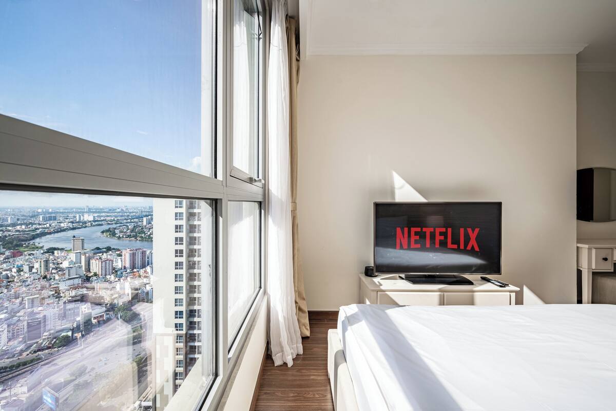 Habitación de hotel con un ventanal grande, una cama, un televisor mostrando 'NETFLIX' y vista a la ciudad.