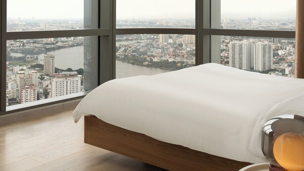 Un dormitorio con una cama grande, una mesita de noche y vistas a un paisaje urbano a través de amplios ventanales.