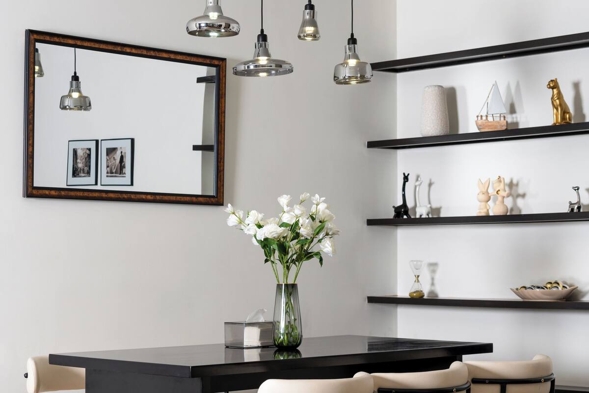 Un moderno comedor con una mesa negra, seis sillas, un jarrón de flores blancas, luces colgantes, un espejo y estanterías con adornos.