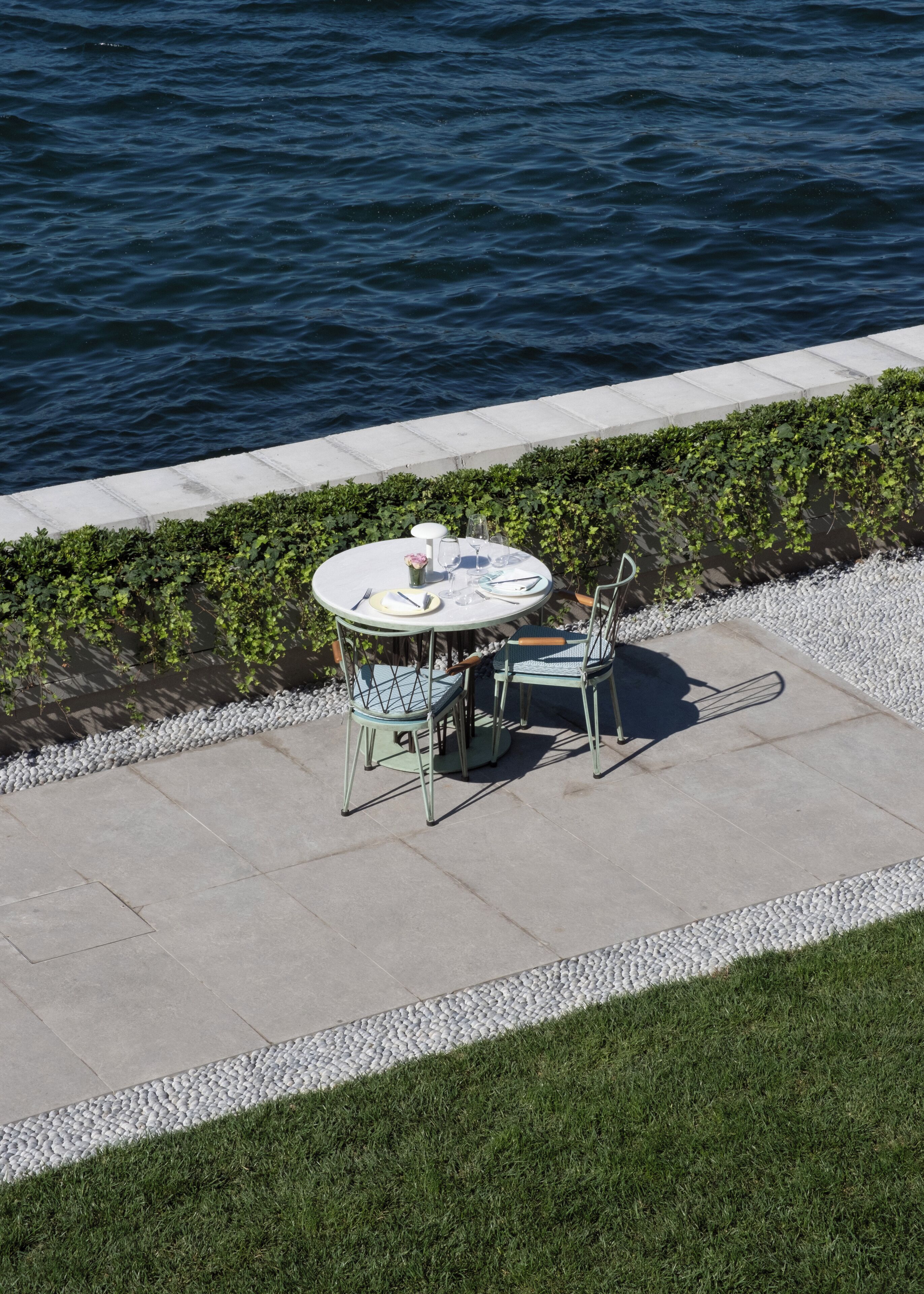 A round table with chairs and a small vase of flowers, set on a concrete patio by a body of water.