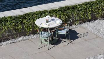 A round table with chairs and a small vase of flowers, set on a concrete patio by a body of water.