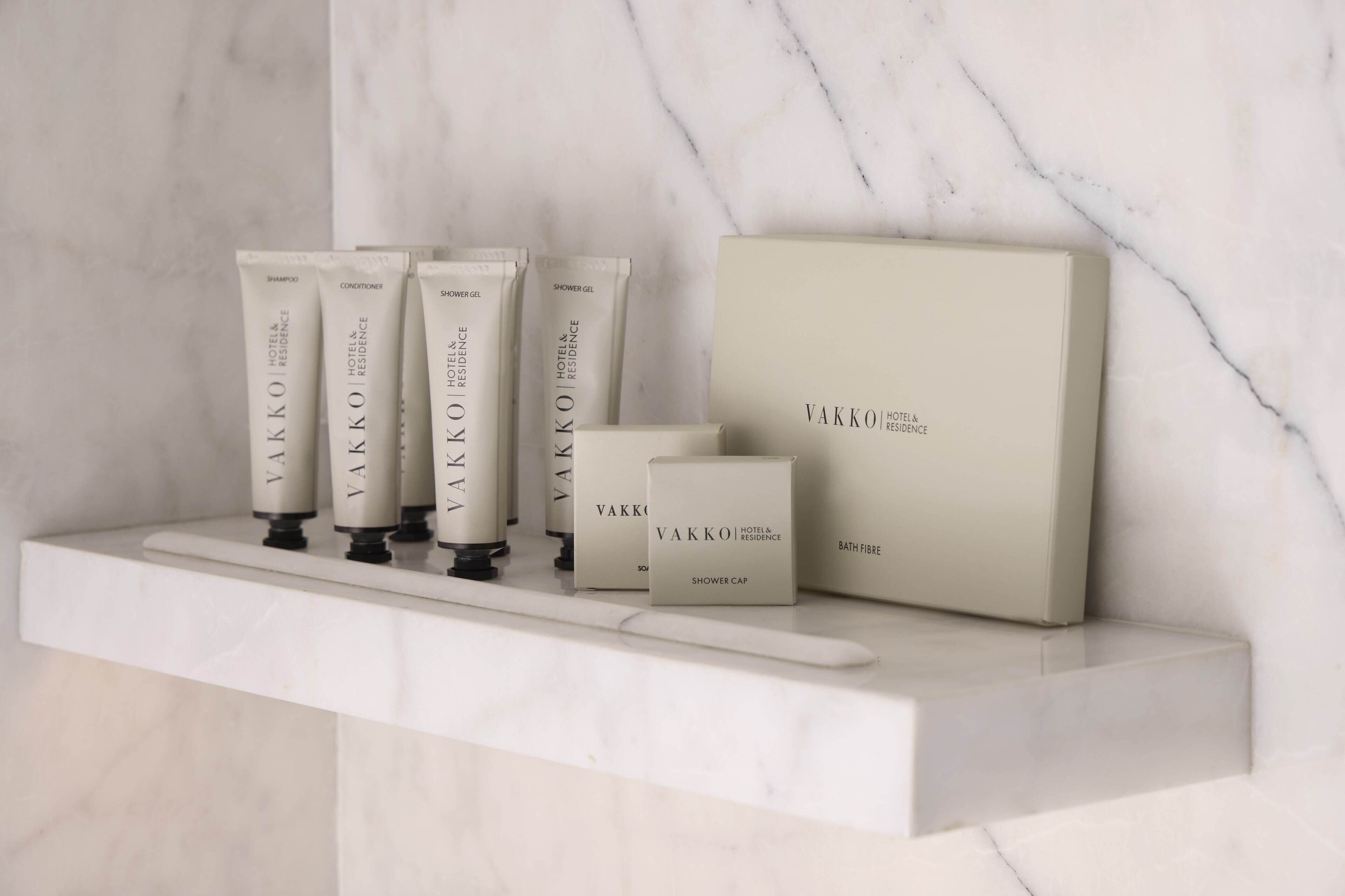 A marble shelf with VAKKO hotel & residence toiletries including shampoo, conditioner, shower gel, and a shower cap.