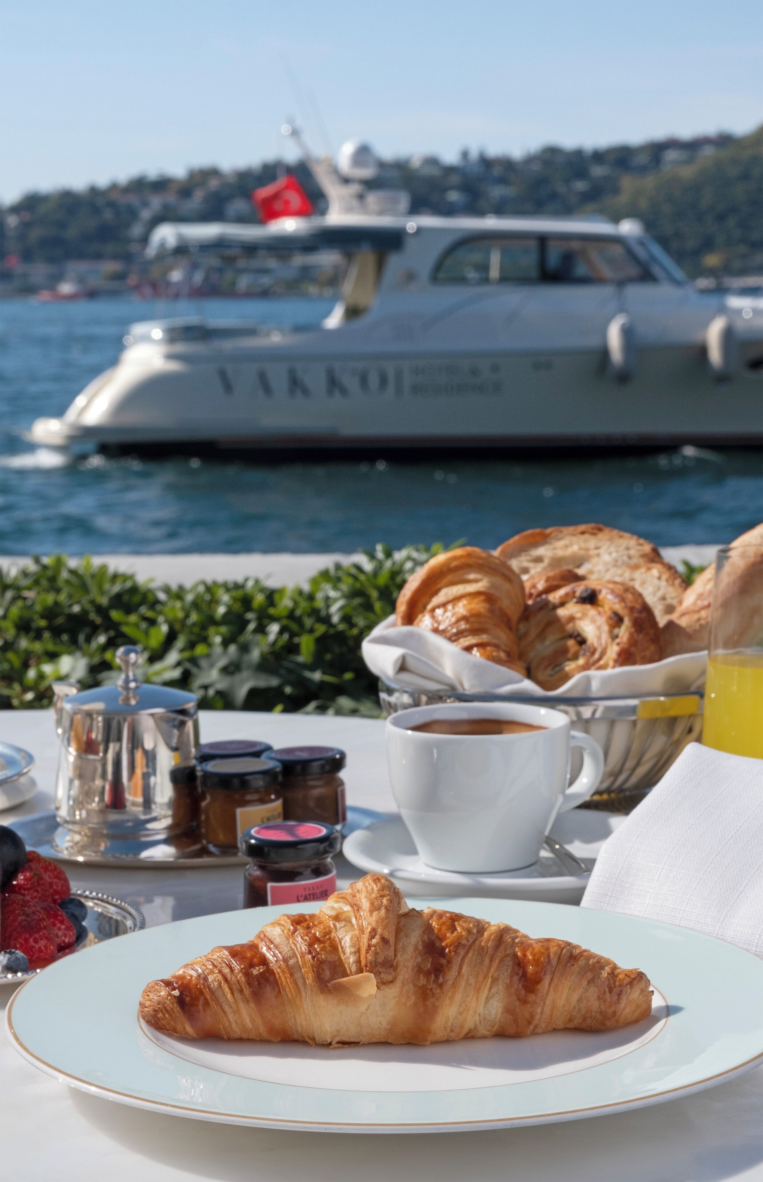 A croissant on a plate, a cup of coffee, and a glass of orange juice on a table with a view of a boat in the background.