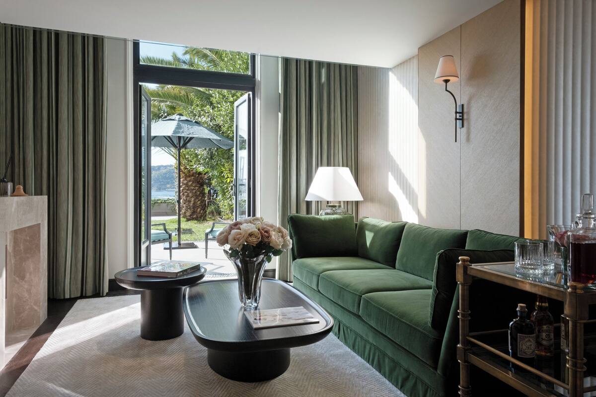 Ein modernes Hotelzimmer mit einem grünen Sofa, einem Couchtisch mit Blumenvase, einer Lampe und Blick ins Freie.