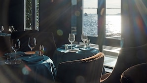 Ein Essbereich mit Tischen für zwei Personen, große Fenster mit Meerblick und eine helle Lichtquelle, die Streulicht erzeugt.
