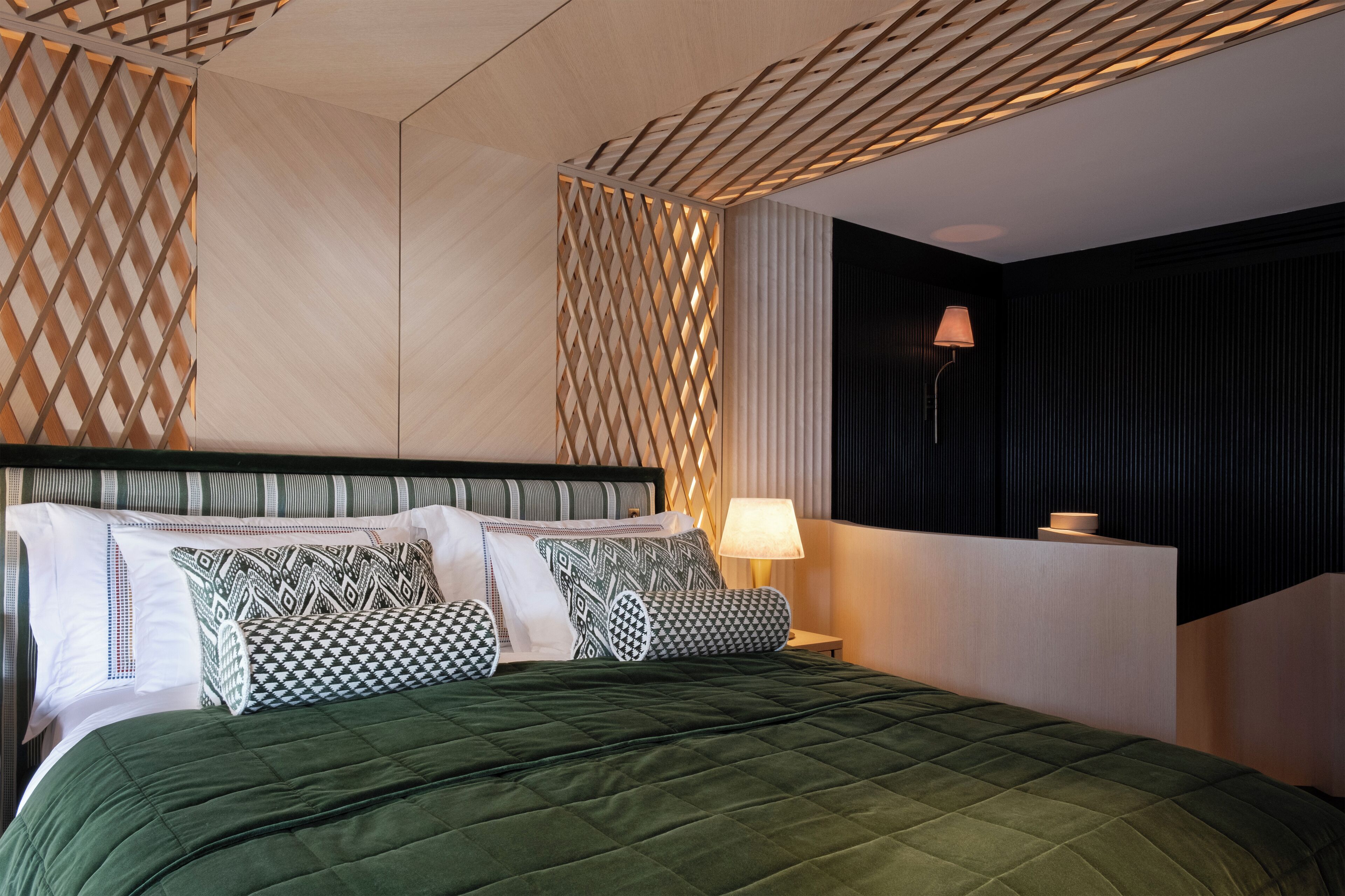A neatly made bed with patterned pillows, a bedside lamp, and a wall with a geometric design.