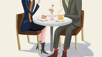 A man and a woman sitting at a round table with a vase of flowers, plates of food, and a glass of red drink.