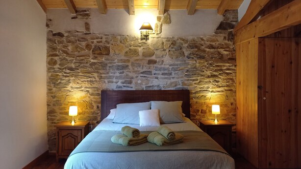 A bedroom with a stone wall, wooden ceiling beams, a wooden headboard, and two bedside lamps.