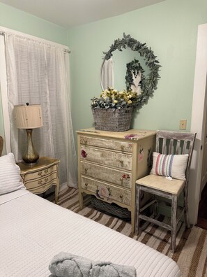 A bedroom with a bed, a dresser, a chair, a lamp, a basket with flowers, and a mirror.