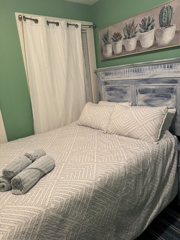 A neatly made bed with a patterned bedspread and pillows, a window with white curtains, and a wall-mounted painting of potted plants.
