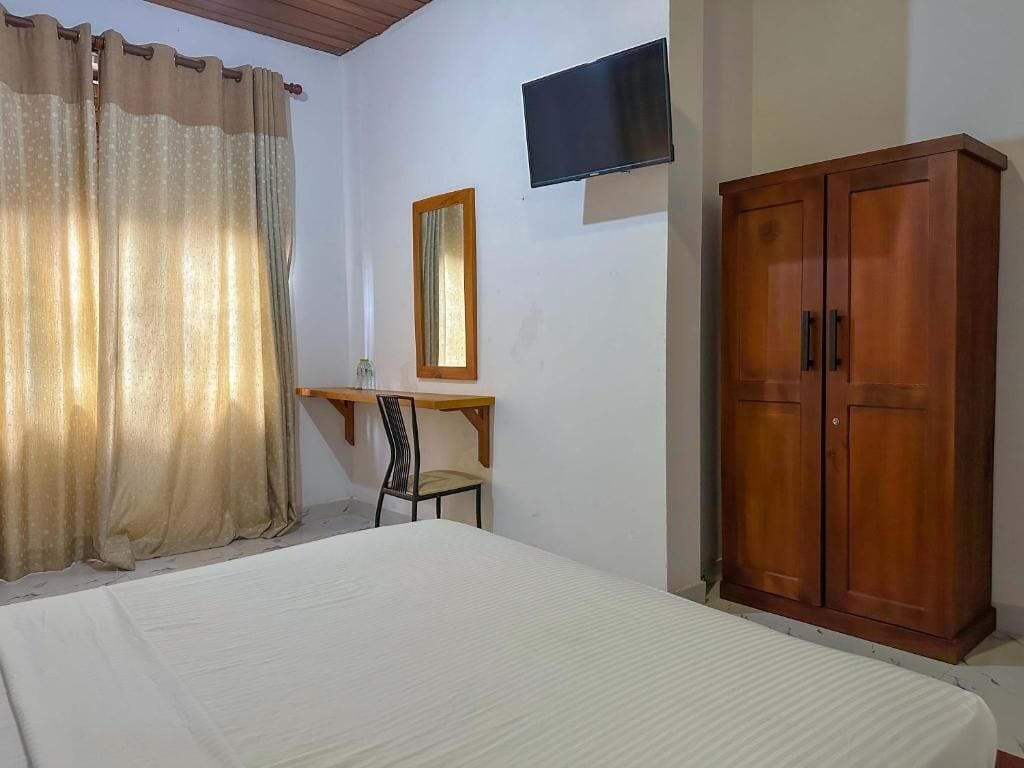A bedroom with a bed, a wooden wardrobe, a television mounted on the wall, and a desk with a chair.