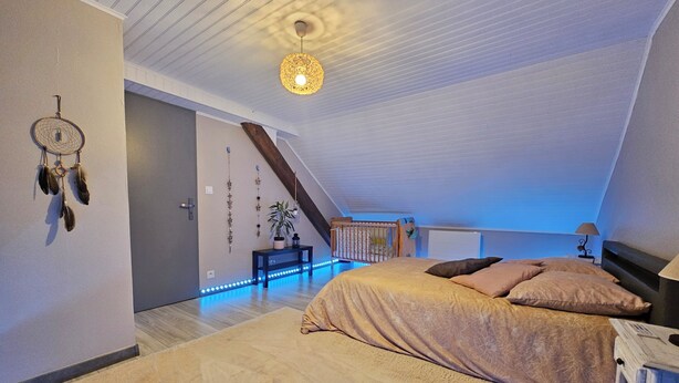 Une chambre avec un plafond en pente, un lit recouvert d’une couverture beige, un attrape-rêves accroché au mur et une plante suspendue.