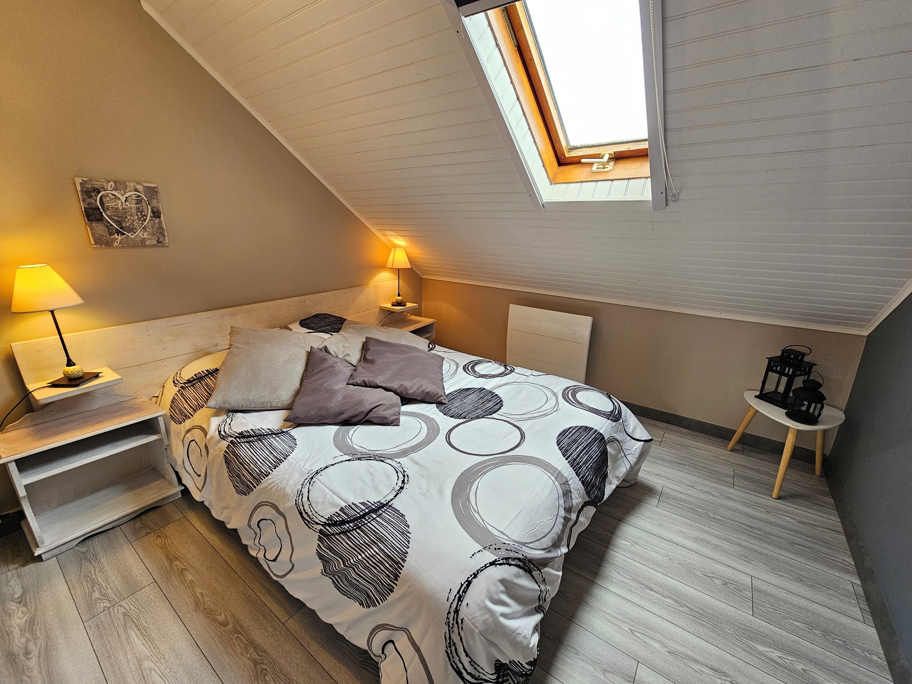 A bedroom with a sloped ceiling, a skylight, a bed with patterned bedding, bedside lamps, and a small table with a lantern.