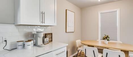 Una cocina moderna con armarios blancos, cafetera y un comedor con mesa y sillas de madera.