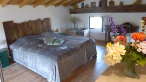 Un dormitorio con techo de madera, una cama con colcha gris, un aparador, una ventana y un jarrón con flores.