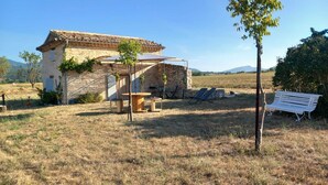 Un edificio rústico de piedra con techo de tejas, rodeado por un campo cubierto de hierba y algunos árboles. Hay zonas de estar al aire libre y un banco.