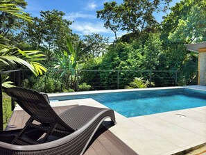 Una piscina con una tumbona de mimbre al frente, rodeada de abundante vegetación.