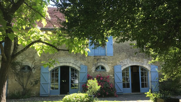 Un bâtiment rustique en pierre, doté de fenêtres en arc de cercle et de volets bleus, entouré d’une végétation luxuriante et de plantes en fleurs.