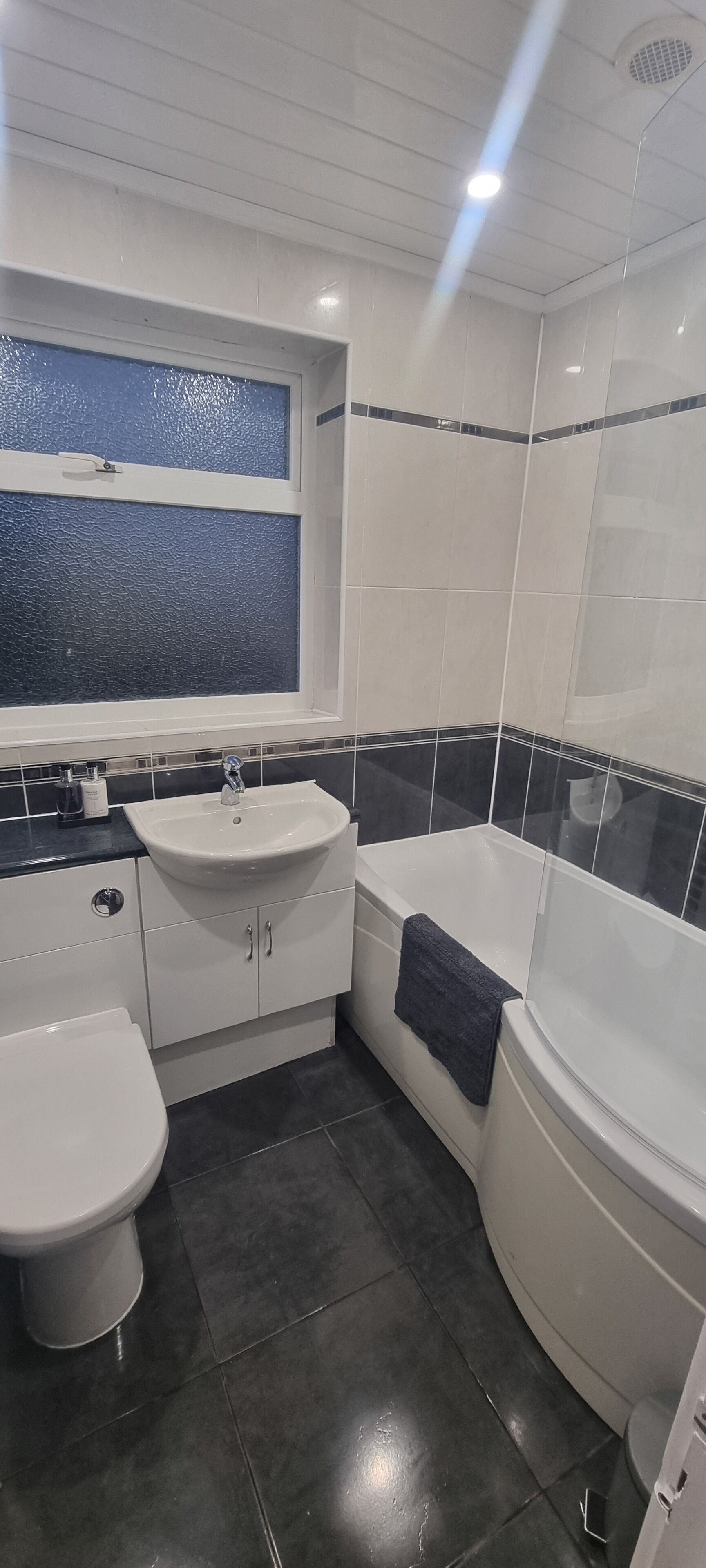 A bathroom with a white sink, toilet, and bathtub, black and white tiles, and a window with a textured glass panel.