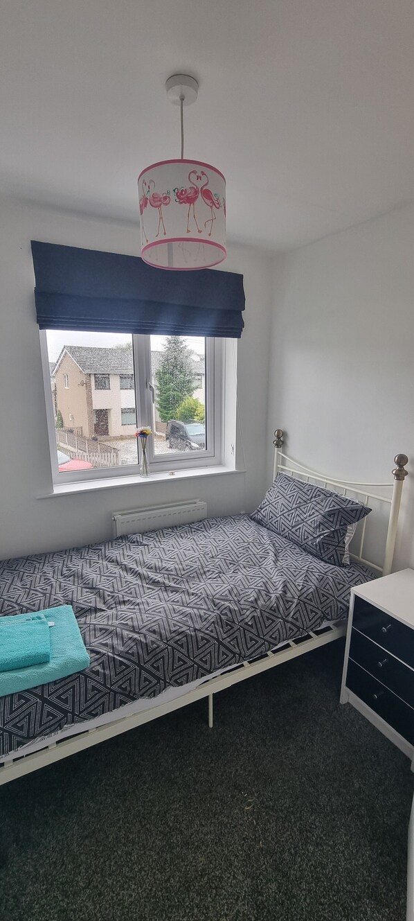 Una cama individual con una colcha de estampado geométrico, una ventana con vistas a casas, un radiador blanco y una mesita de noche con cajones.