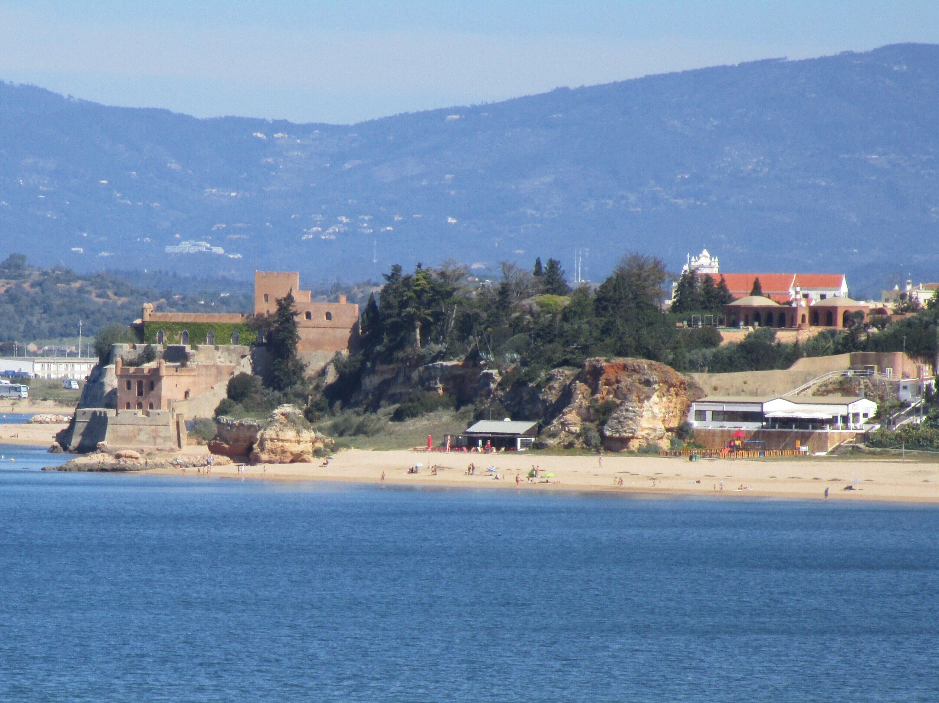A coastal town with a historic fort, beach, and a hilltop building with a red roof.