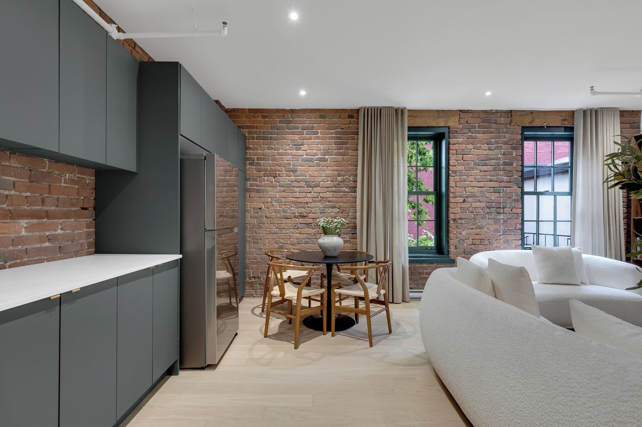 A modern kitchen with grey cabinets, a brick wall, a dining area with a round table and chairs, and a white sofa.
