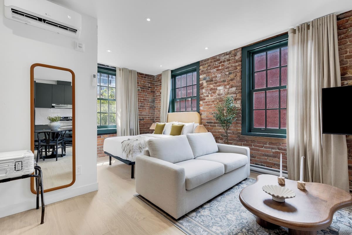 Ein modernes Wohnzimmer mit einem weißen Sofa, einem Holztisch und einem großen Spiegel. Ein Bett mit einem grünen Kopfteil, ein an der Wand montierter Fernseher und eine Ziegelwand setzen Akzente im Raumdesign.