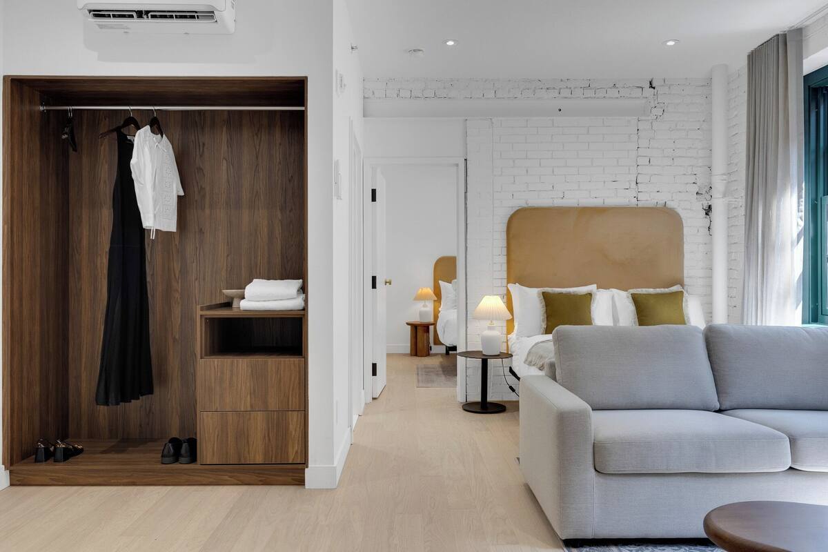 Ein modernes Hotelzimmer mit einem Holzschrank, einem grauen Sofa, einem Bett mit weißem Kopfteil und einem kleinen Tisch mit Lampe.