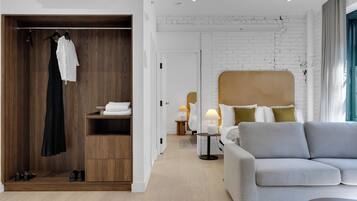 Una habitación de hotel moderna con un armario de madera, un sofá gris, una cama con cabecero blanco y una mesita de noche con lámpara.