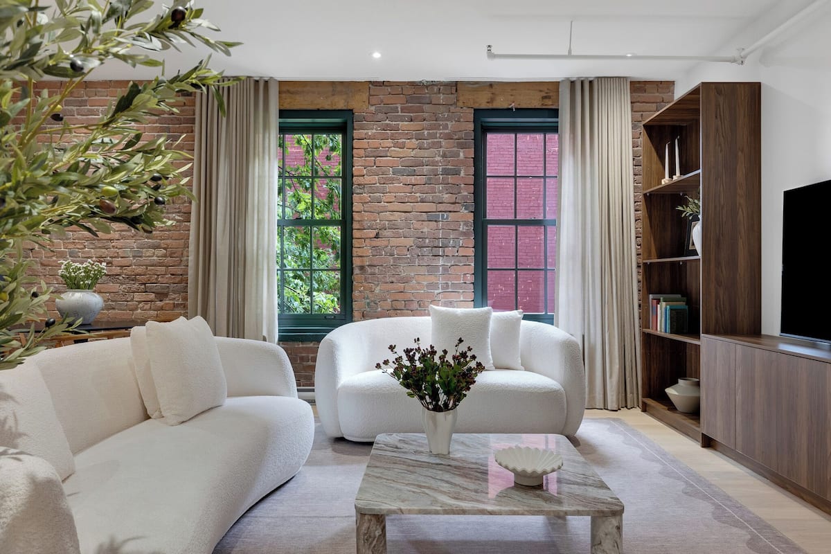 Ein modernes Wohnzimmer mit Backsteinwand, zwei mit Vorhängen versehenen Fenstern, einem weißen Sofa, einem Couchtisch mit einer Vase und einem Holzregal.
