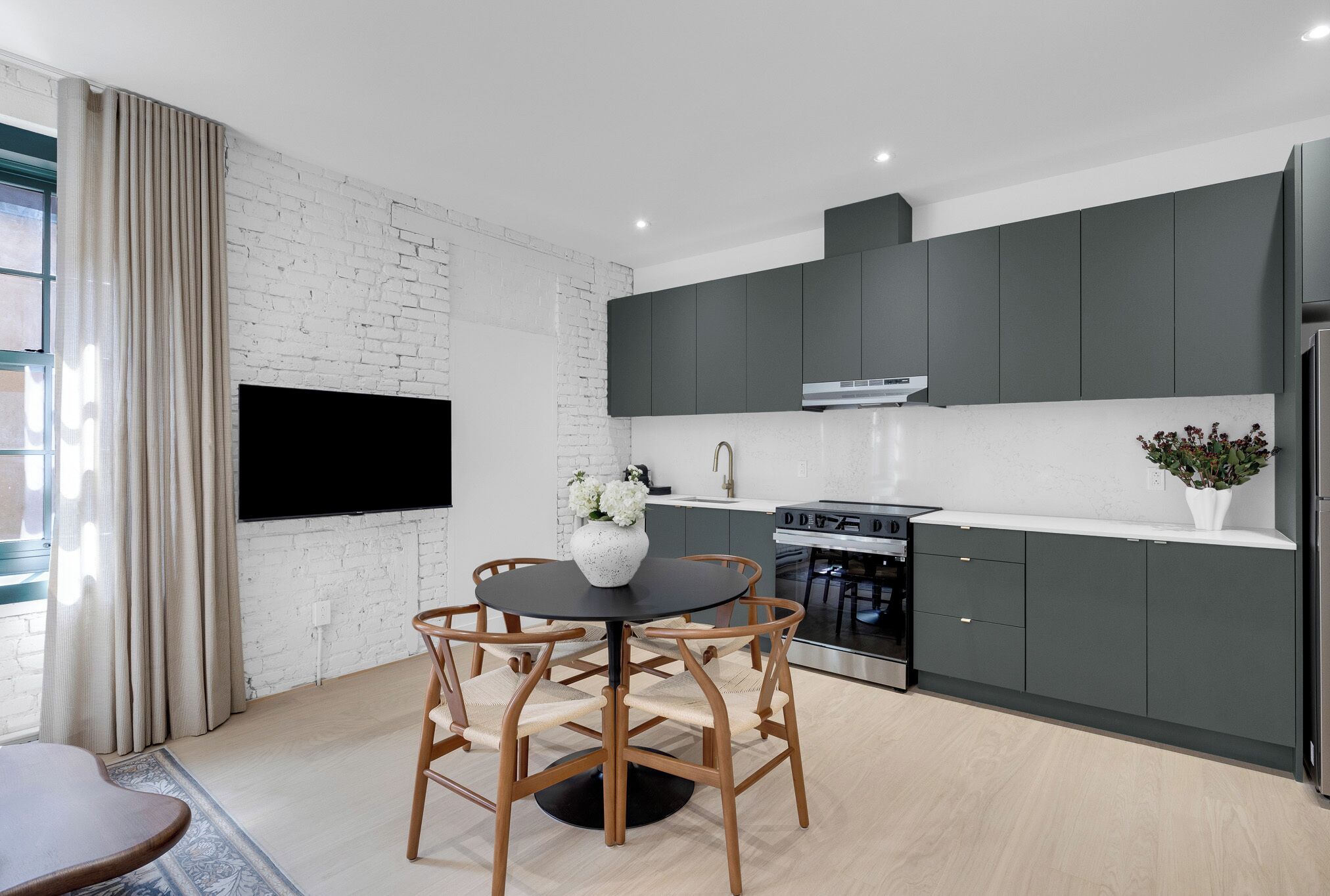 A modern kitchen with dark cabinets, a black dining table, and a flat-screen TV mounted on the wall.