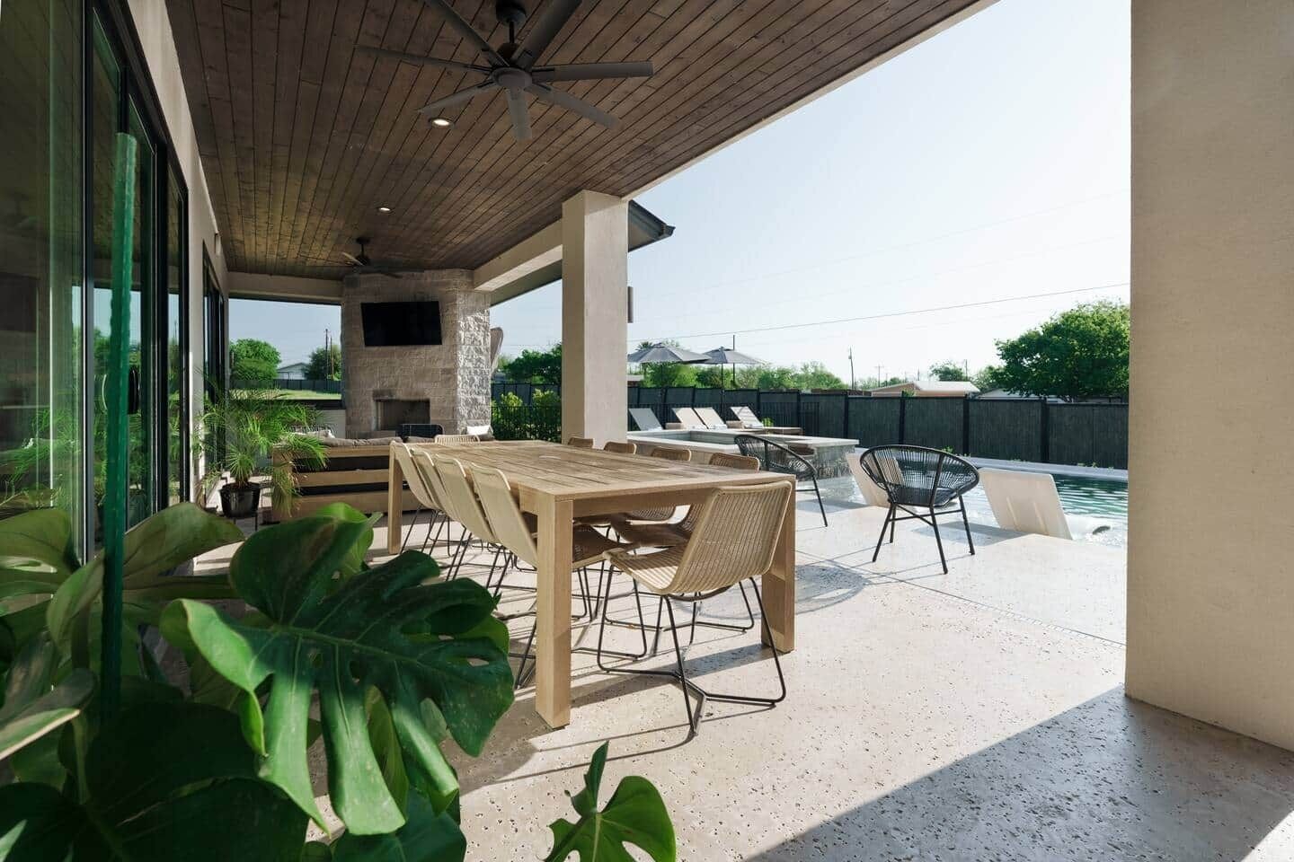 An outdoor patio with a wooden table and chairs, a stone fireplace, and a pool area.