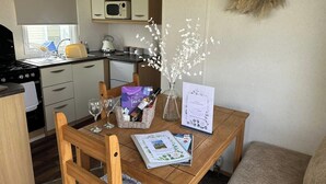 A compact kitchen with a microwave, stove, and sink. A dining table with a placemat, wine glasses, and a bottle of wine is set. A vase with white flowers and a menu are also on the table. - Marine Getaways Caravan Hire

A home from home experience in luxury.. (Rhyl)