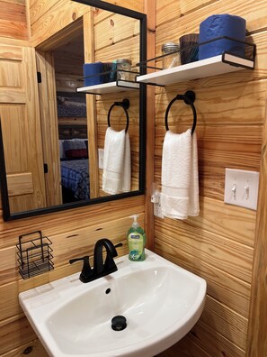 Une salle de bain de style rustique, avec des murs en bois, un lavabo blanc, un robinet noir et un miroir au-dessus. Deux serviettes sont suspendues à des porte-serviettes noirs, et une étagère contient des pots et un récipient bleu.