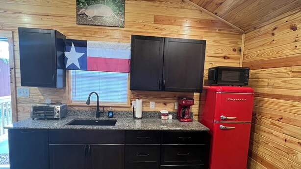 A compact kitchen with wooden walls, a black countertop, a red refrigerator, and a black couch in the foreground. - Cabin Unit 3 of 6 Called the Texan with WiFi and AC in serene Paige (Paige)